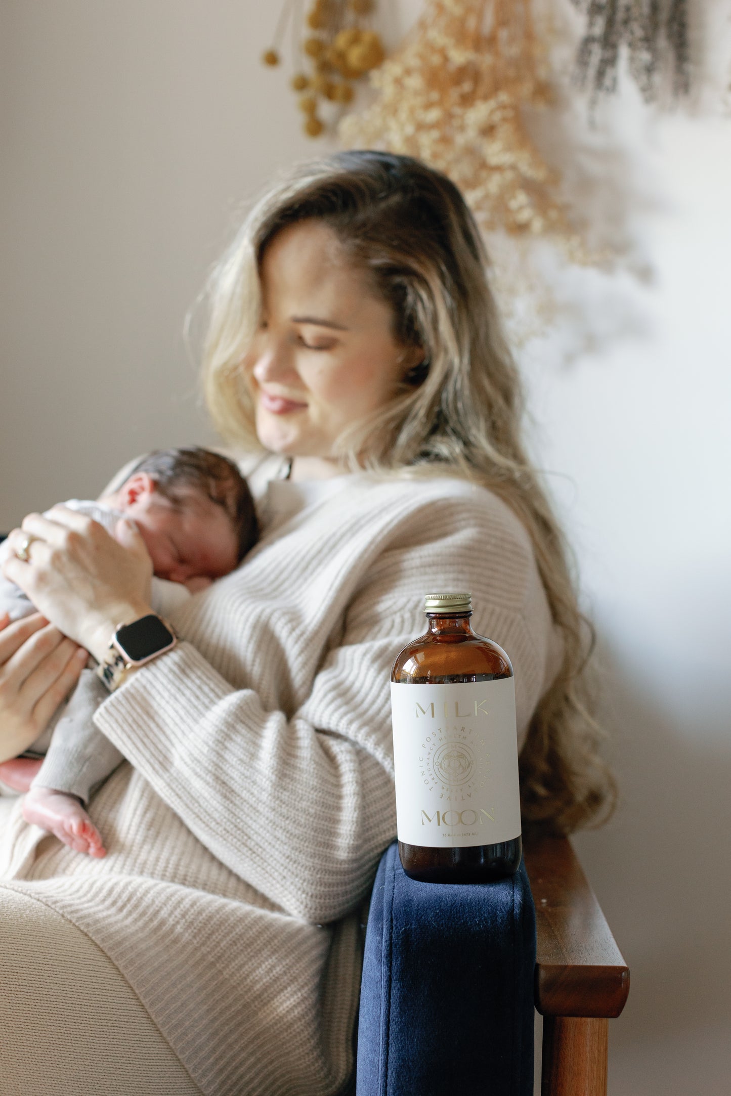 picture of a mother and her baby sitting on a chair with the postpartum restorative tonic with a bottle that has the phrase "Milk Moon" written on it