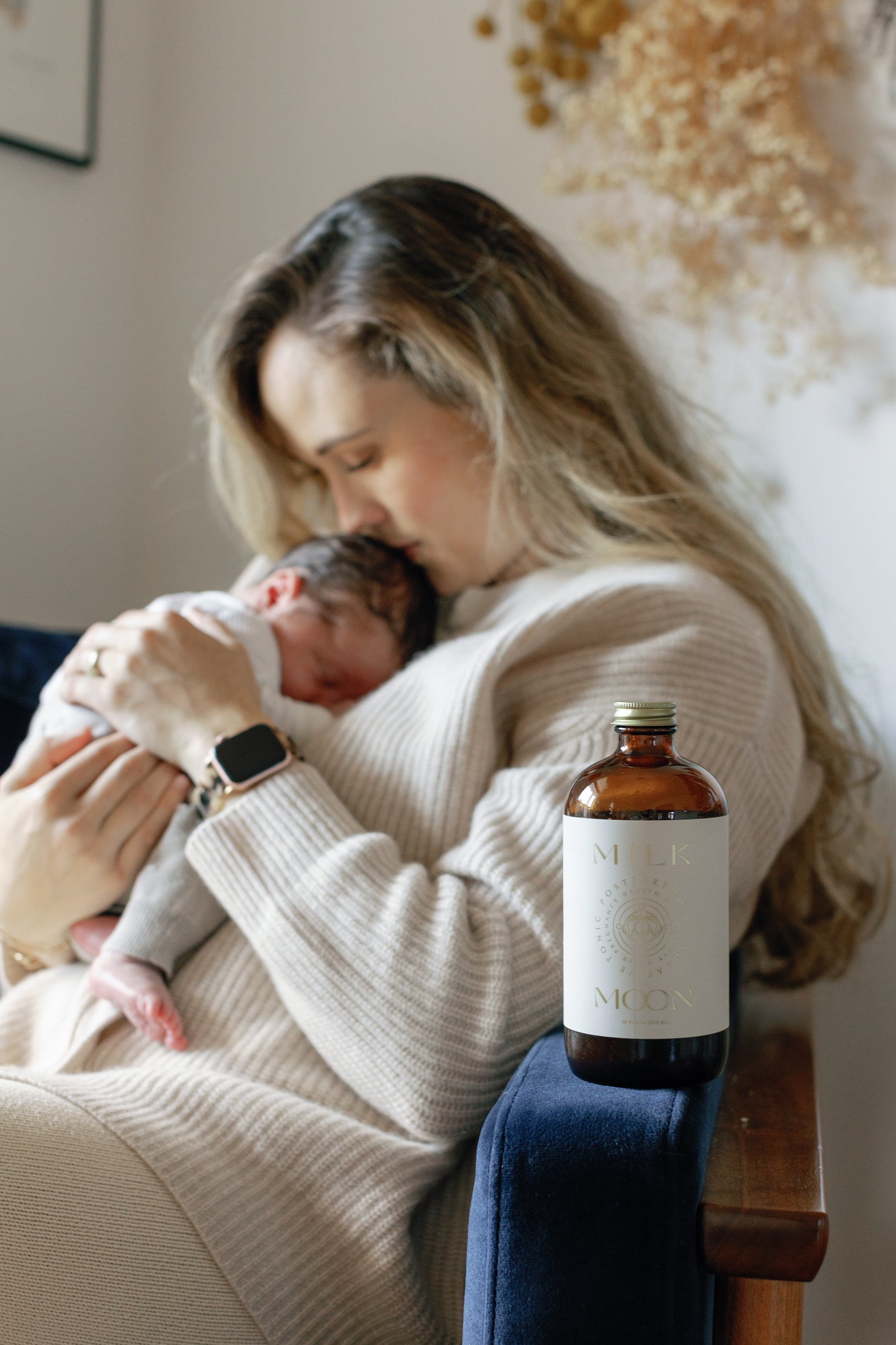 picture of a mother and her baby sitting on a chair with the postpartum restorative tonic with a bottle that has the phrase "Milk Moon" written on it