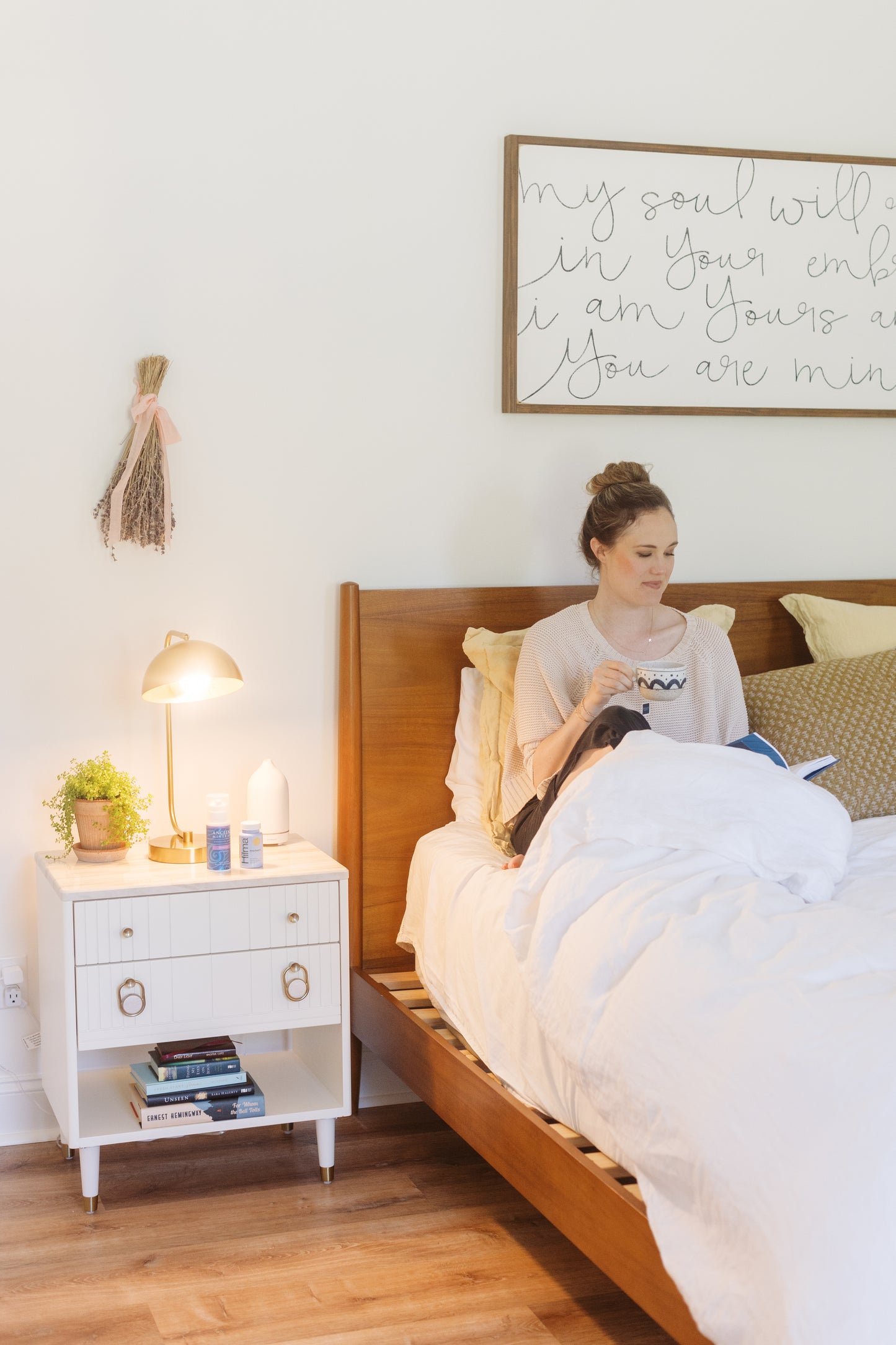 picture of a happy woman sitting on her bed while holding a cup of tea on one hand and on the other one a book